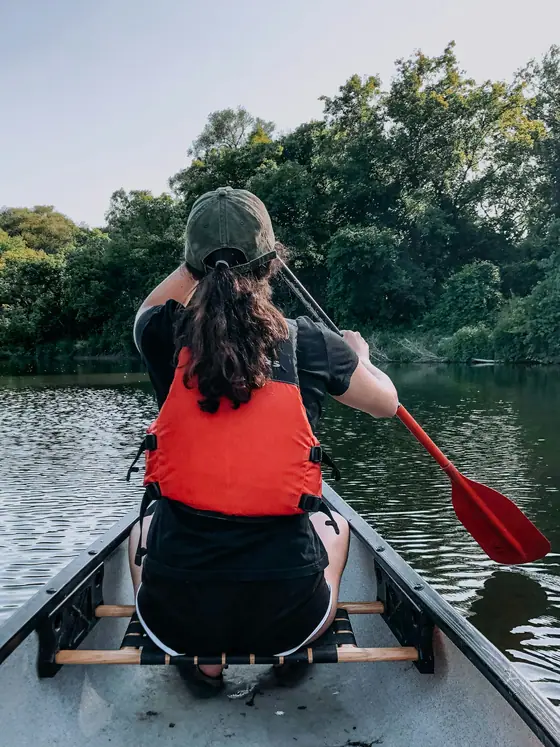 Une pagayeuse en gilet de sauvetage orange, de dos, avance dans un canoë le long d'une rive boisée